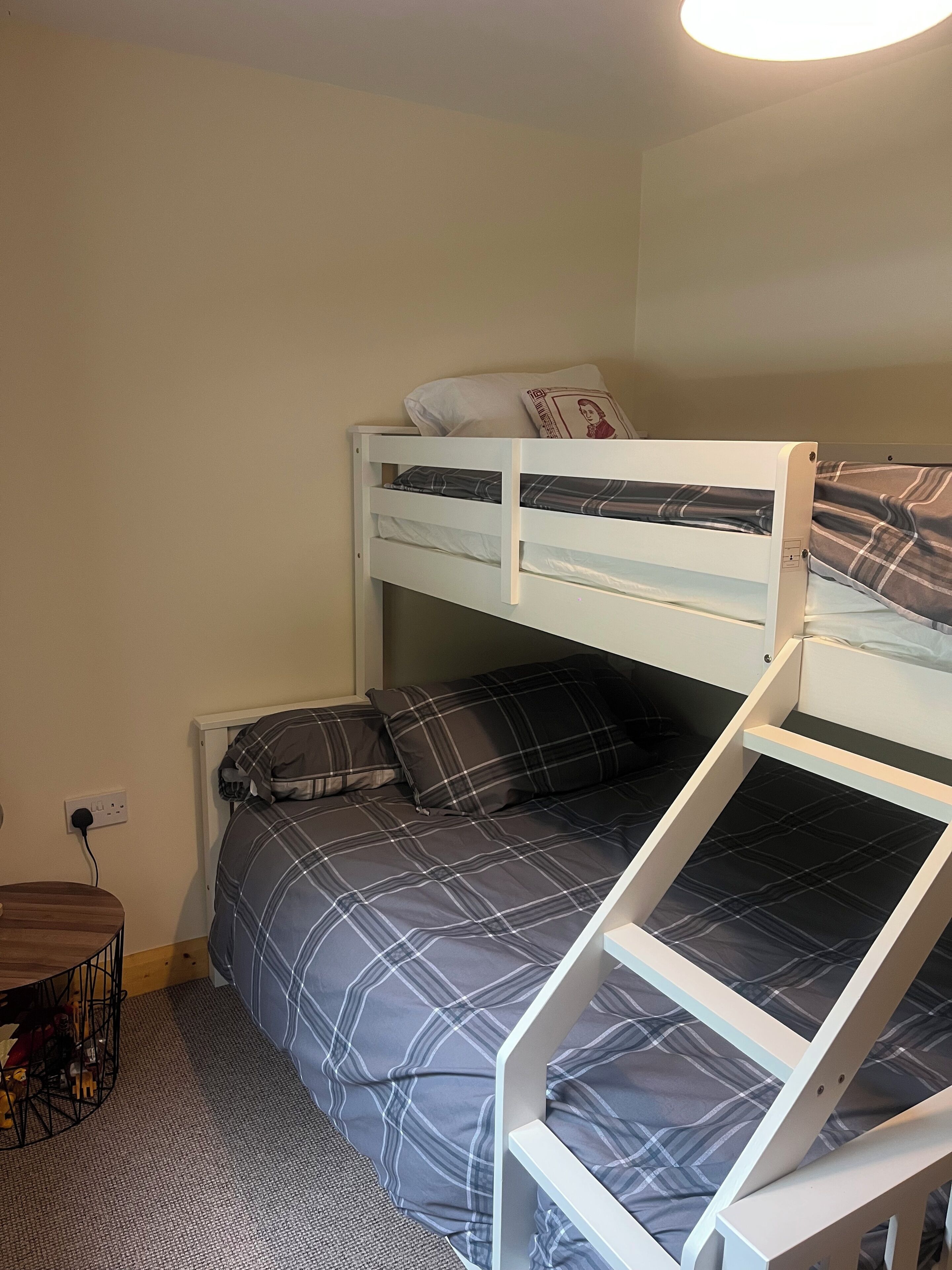 A bunk bed with a lower bed and a higher bunk, both with plaid bedding. There is a small wooden table with a metal frame and a power outlet on the wall.