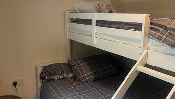 A bunk bed with a lower bed and a higher bunk, both with plaid bedding. There is a small wooden table with a metal frame and a power outlet on the wall.
