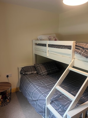 A bunk bed with a lower bed and a higher bunk, both with plaid bedding. There is a small wooden table with a metal frame and a power outlet on the wall.