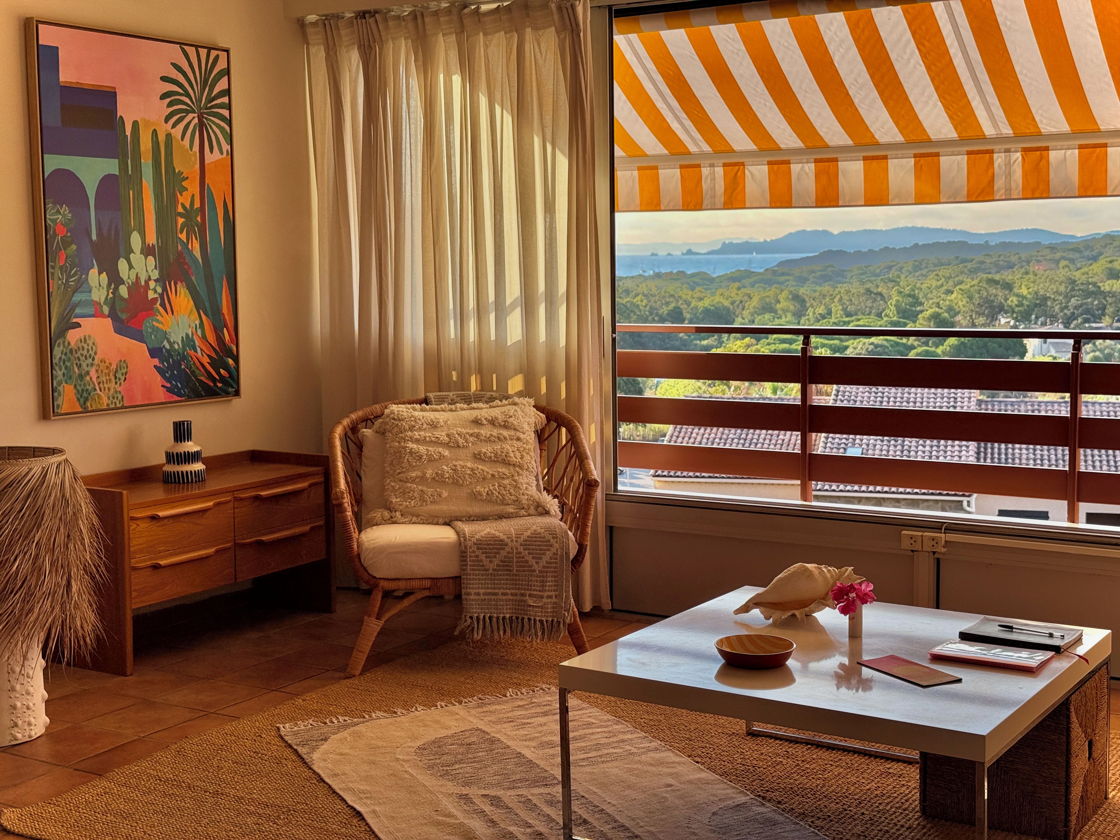 Une pièce offrant une vue sur un paysage montagneux, avec un fauteuil en osier, une table d’appoint en bois et un tableau accroché au mur.
