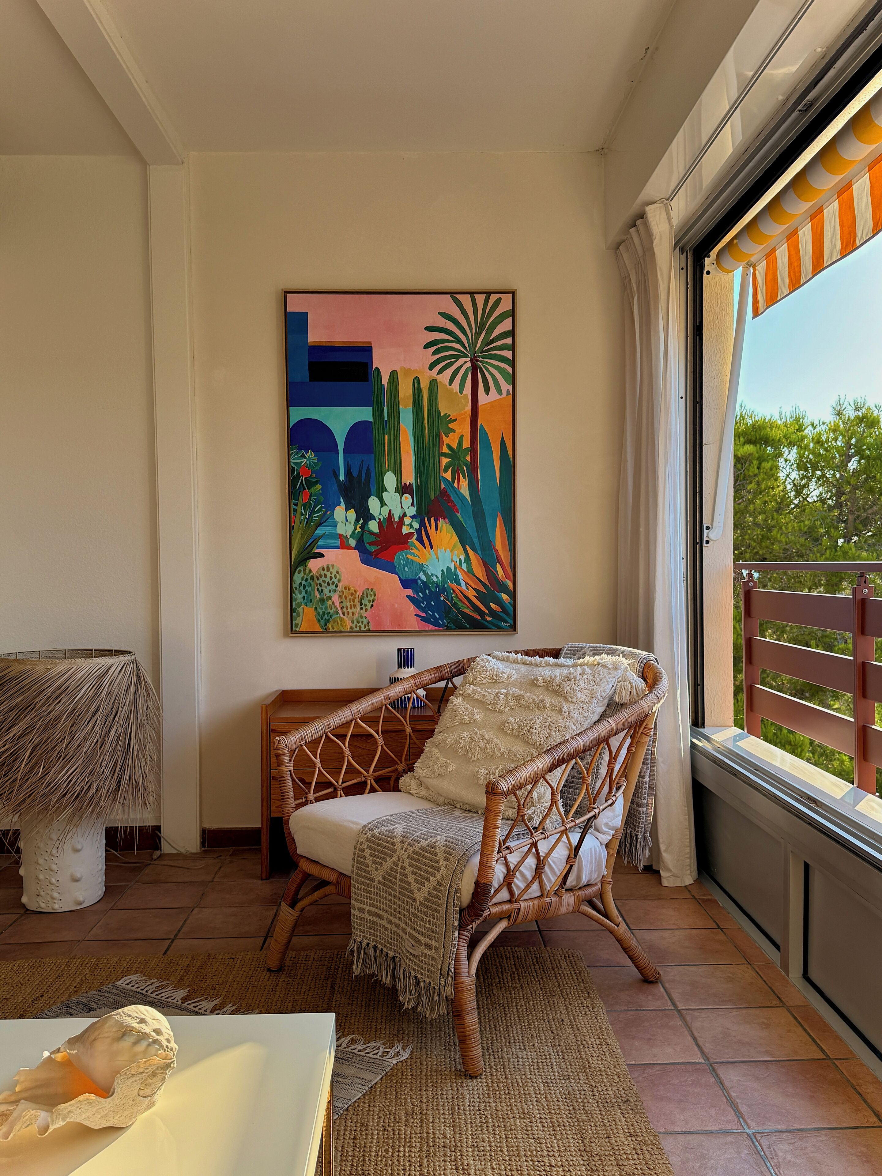 A wicker chair with a throw blanket, a painting of a colorful landscape, and a view of greenery through a window.