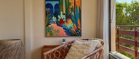 A wicker chair with a throw blanket, a painting of a colorful landscape, and a view of greenery through a window.