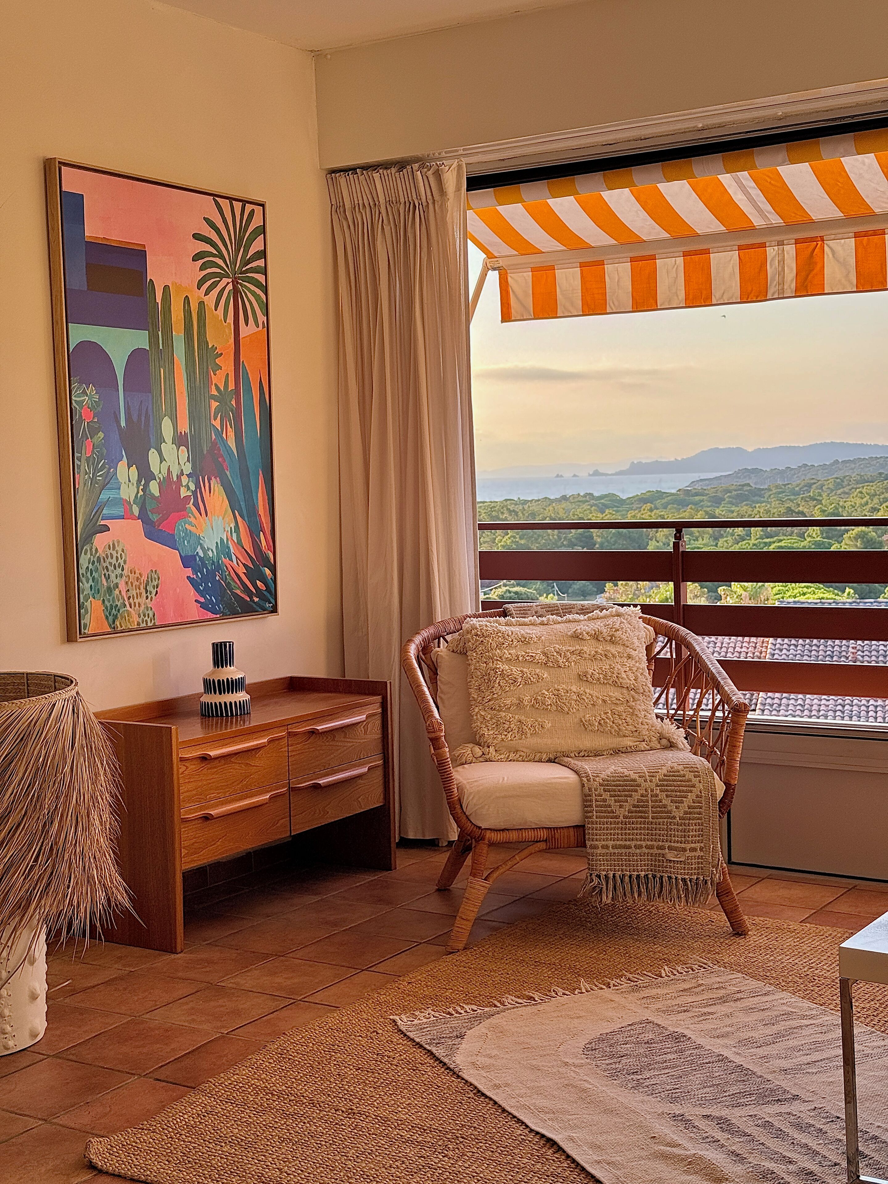 A room with a view of a mountainous landscape, a wicker chair with a cushion, a wooden side table with a vase, and a colorful painting on the wall.