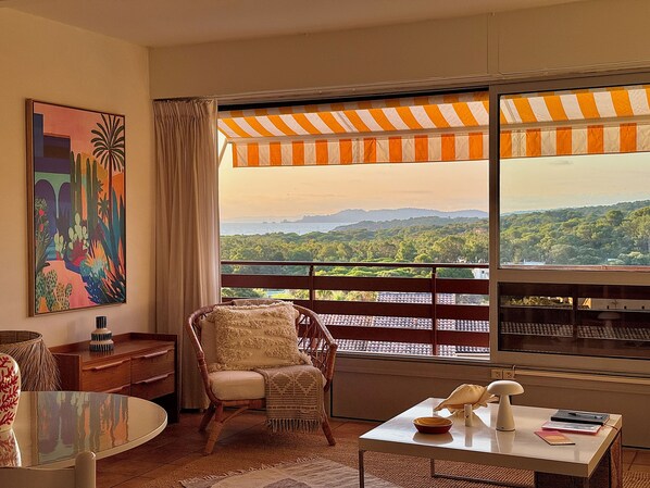 Une pièce avec vue sur un paysage montagneux, une chaise en osier, une table avec un saladier et un tableau accroché au mur.