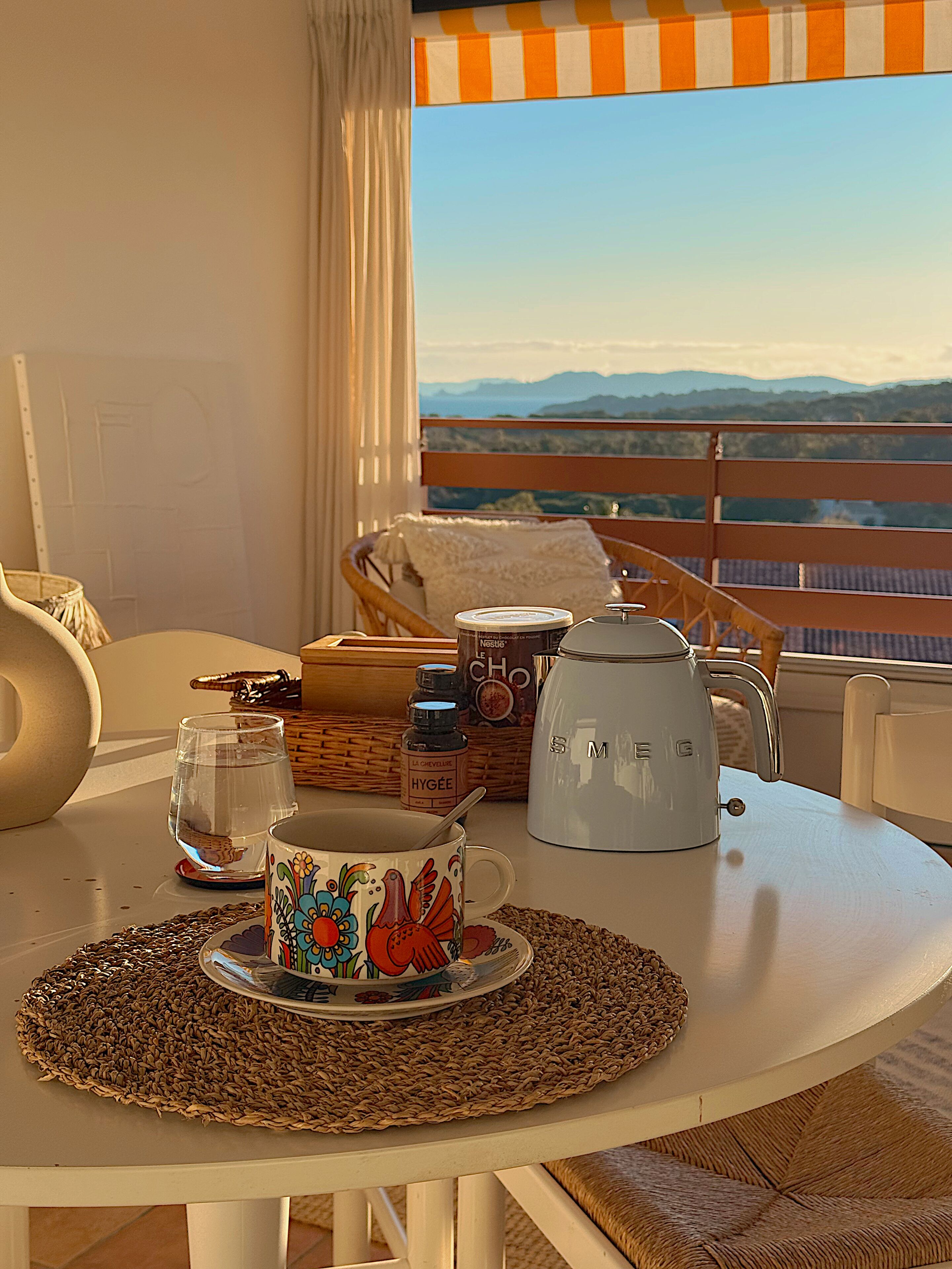A dining table with a coffee pot, a mug, and a bottle of hot chocolate, set on a balcony with a view of mountains.