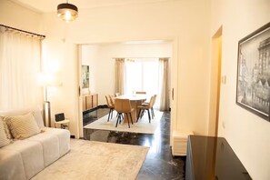 A hotel room with a sofa, a dining table with chairs, a small side table, and a flat-screen TV. - Spacious 4BR Glyfada Retreat Near the Coast (Glifada)