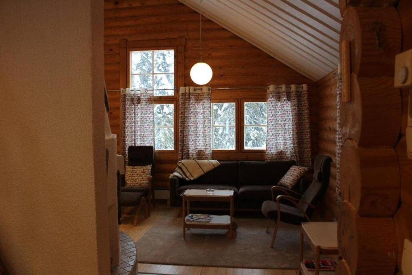 A cozy log cabin interior with a sofa, armchairs, a small table, and a hanging light fixture.