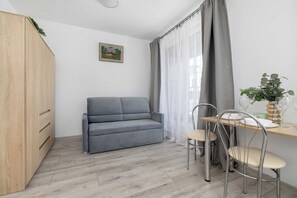 Een kleine woonruimte met een grijze bank, een eettafel met stoelen en een houten garderobe.