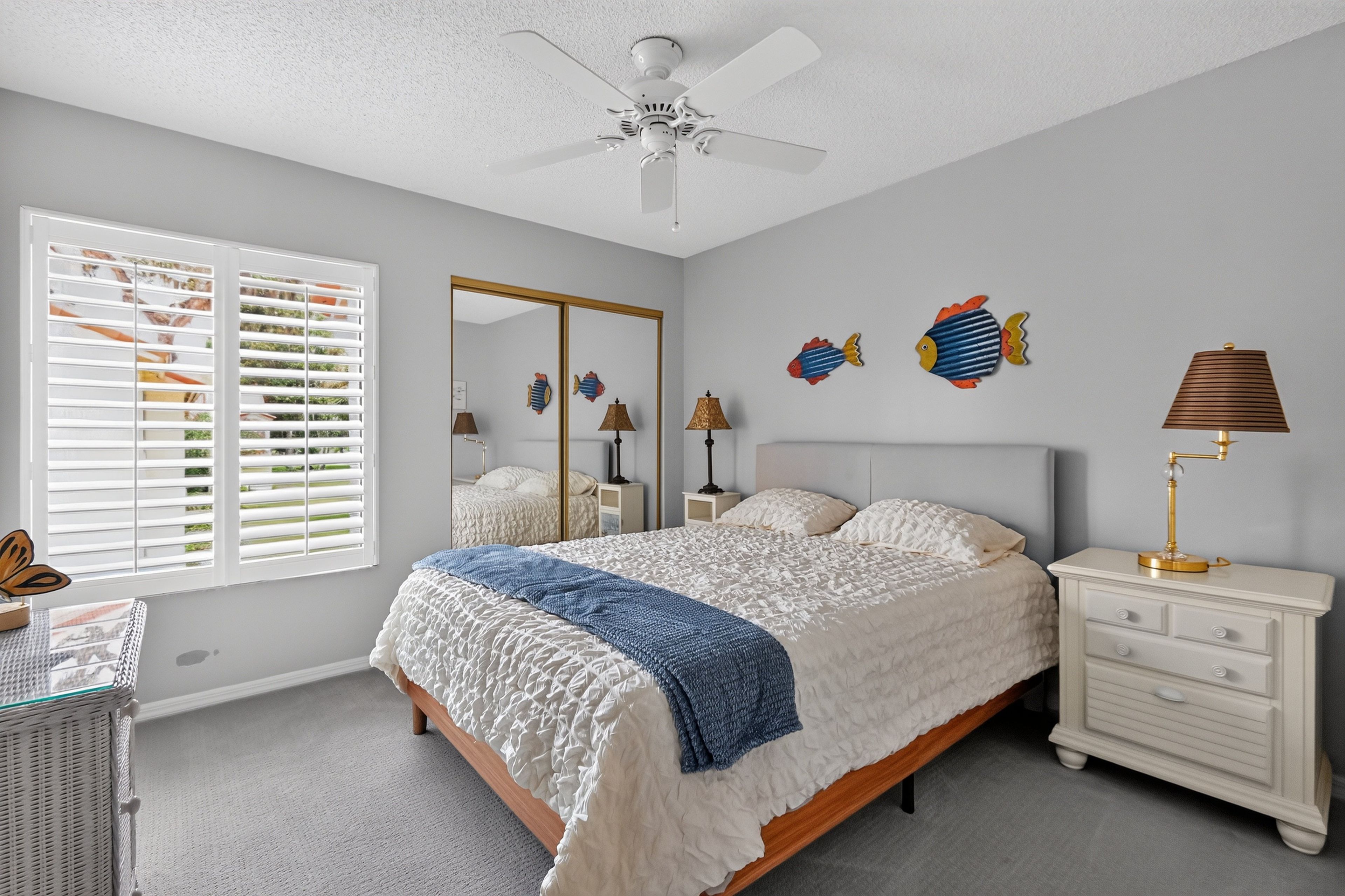 A bedroom with a bed, bedside tables, a ceiling fan, a window with blinds, and wall decorations.