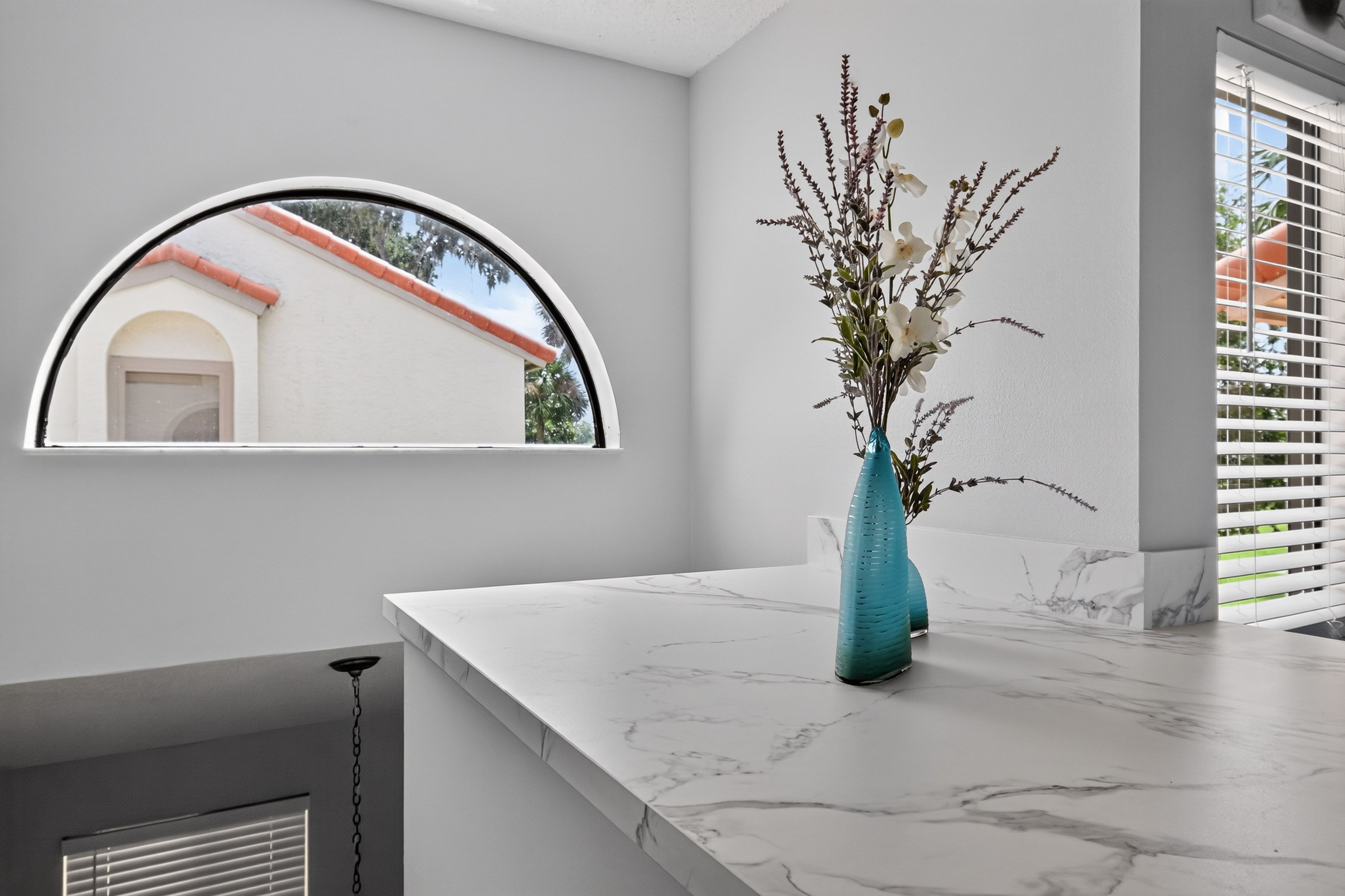 A bathroom with a marble countertop, a vase with flowers, a window with a view of a building, and a ceiling light.