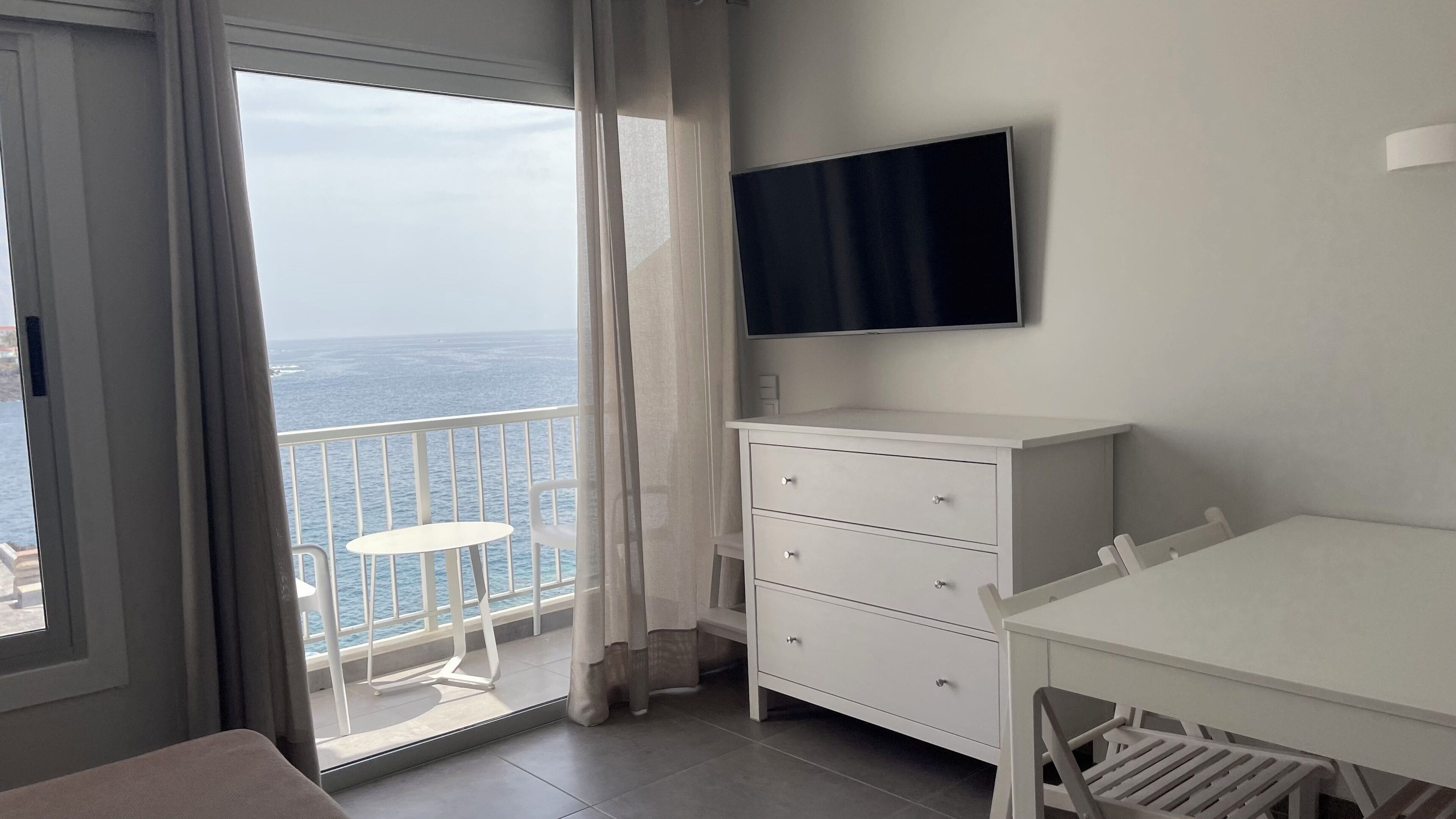 A balcony with a view of the sea, a white dresser, a TV mounted on the wall, and a small table.