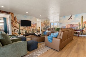 A spacious living room with a large brown sofa, a green armchair, and a coffee table. There is a flat-screen TV mounted on the wall, and the room features a desert-themed mural on the walls. - Red Rock Isle 7 Bedroom Townhouse (Washington)
