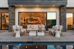 Área de estar à beira da piscina com sofá, mesa de centro, parede de pedra e vista para o interior de uma casa moderna.