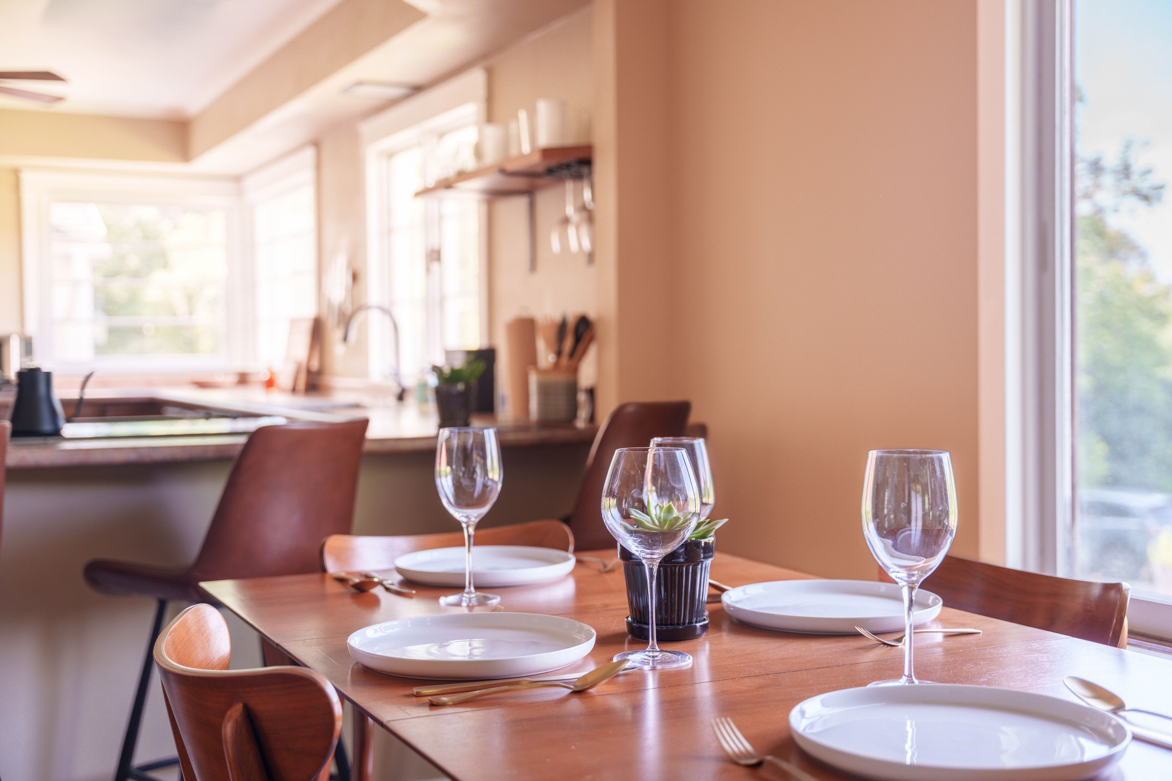 A dining table set with plates, glasses, and cutlery, with a view of the kitchen in the background.