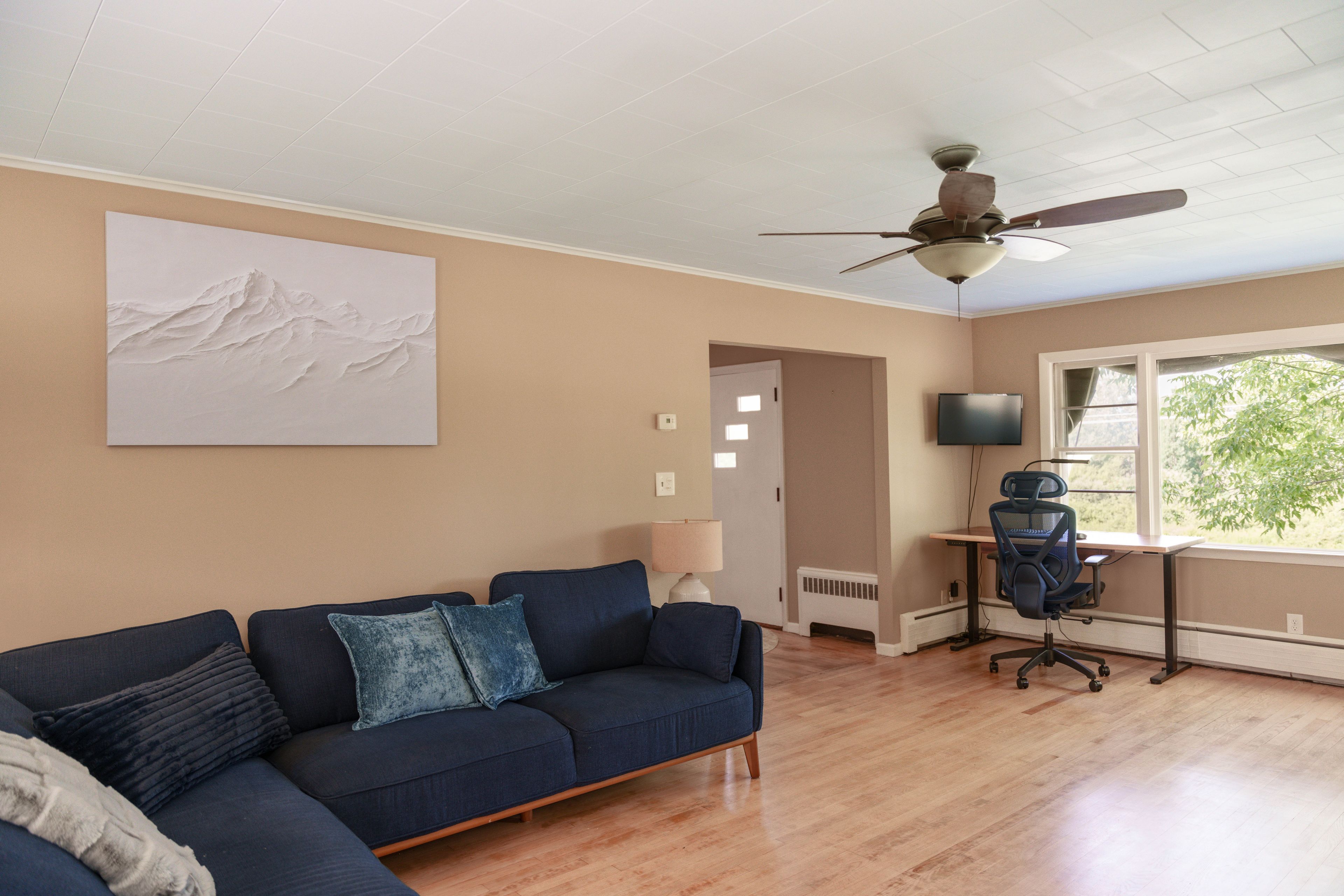 A living room with a sectional sofa, a ceiling fan, a flat-screen TV, and a window with a view of greenery.