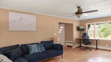 A living room with a sectional sofa, a ceiling fan, a flat-screen TV, and a window with a view of greenery.