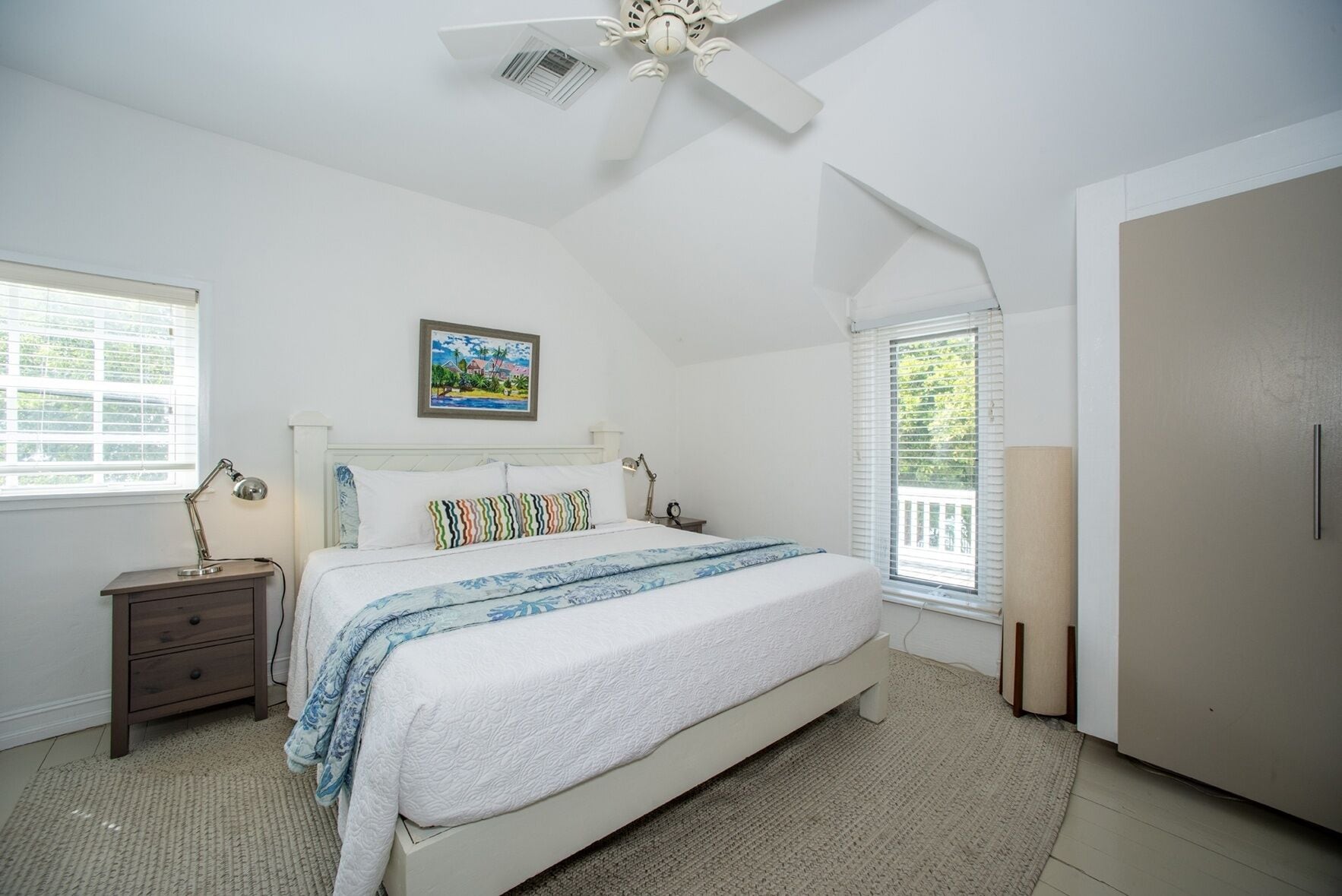 A bedroom with a bed, bedside tables, a ceiling fan, a window with blinds, and a door.