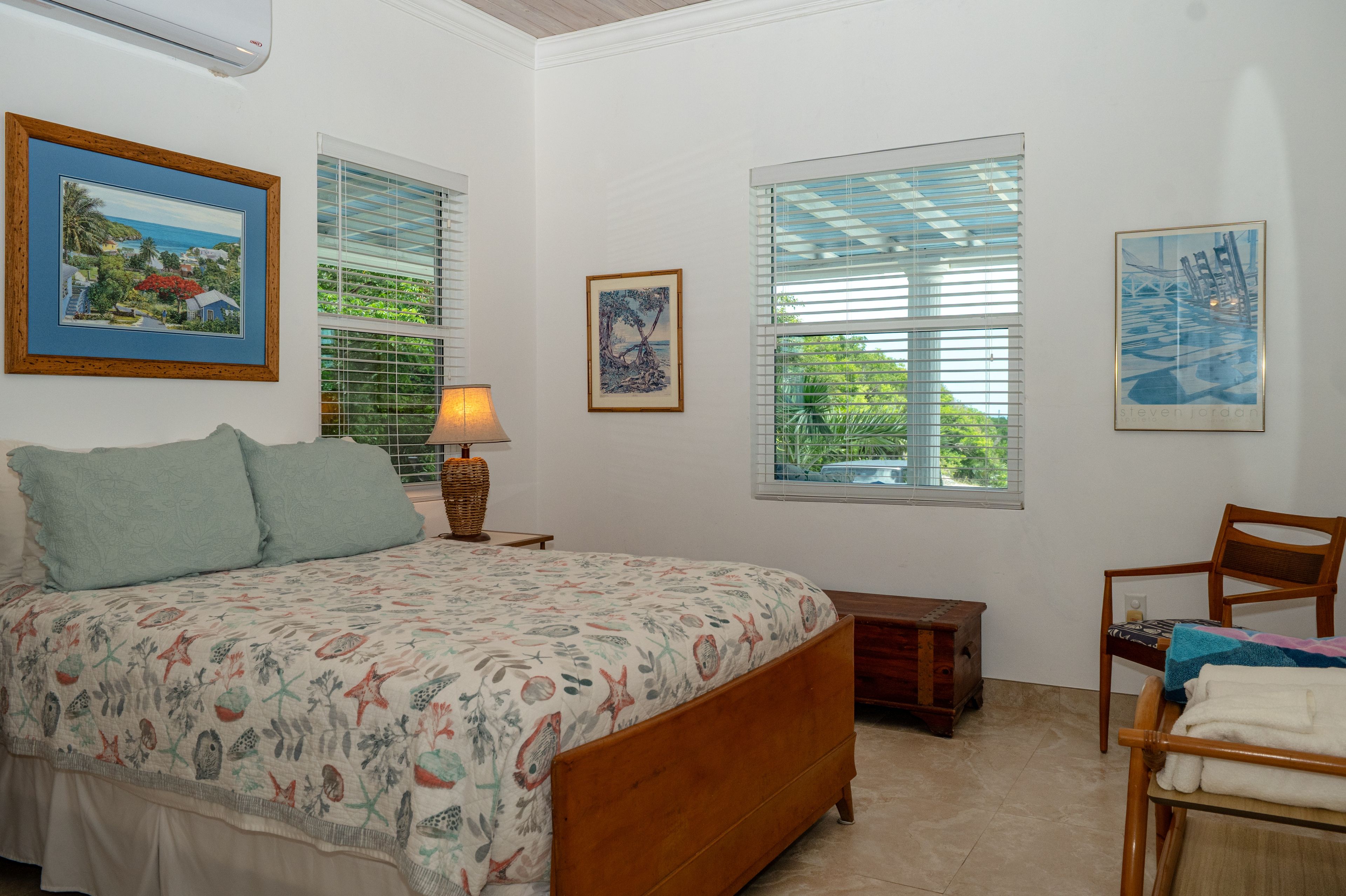 A bedroom with a bed, a nightstand, a lamp, a chair, and two windows with blinds.