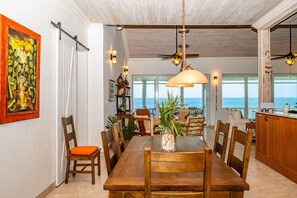 A spacious dining area with a wooden table and chairs, a ceiling fan, and a view of the ocean through large windows. - High Life - Private Hilltop Estate With Pool And Panoramic Ocean 4 Bedroom Home (Governor's Harbour)