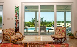 A room with a view of a pool and palm trees, two wicker chairs with floral upholstery, a wooden table, and a decorative wall piece. - High Life - Private Hilltop Estate With Pool And Panoramic Ocean 4 Bedroom Home (Governor's Harbour)
