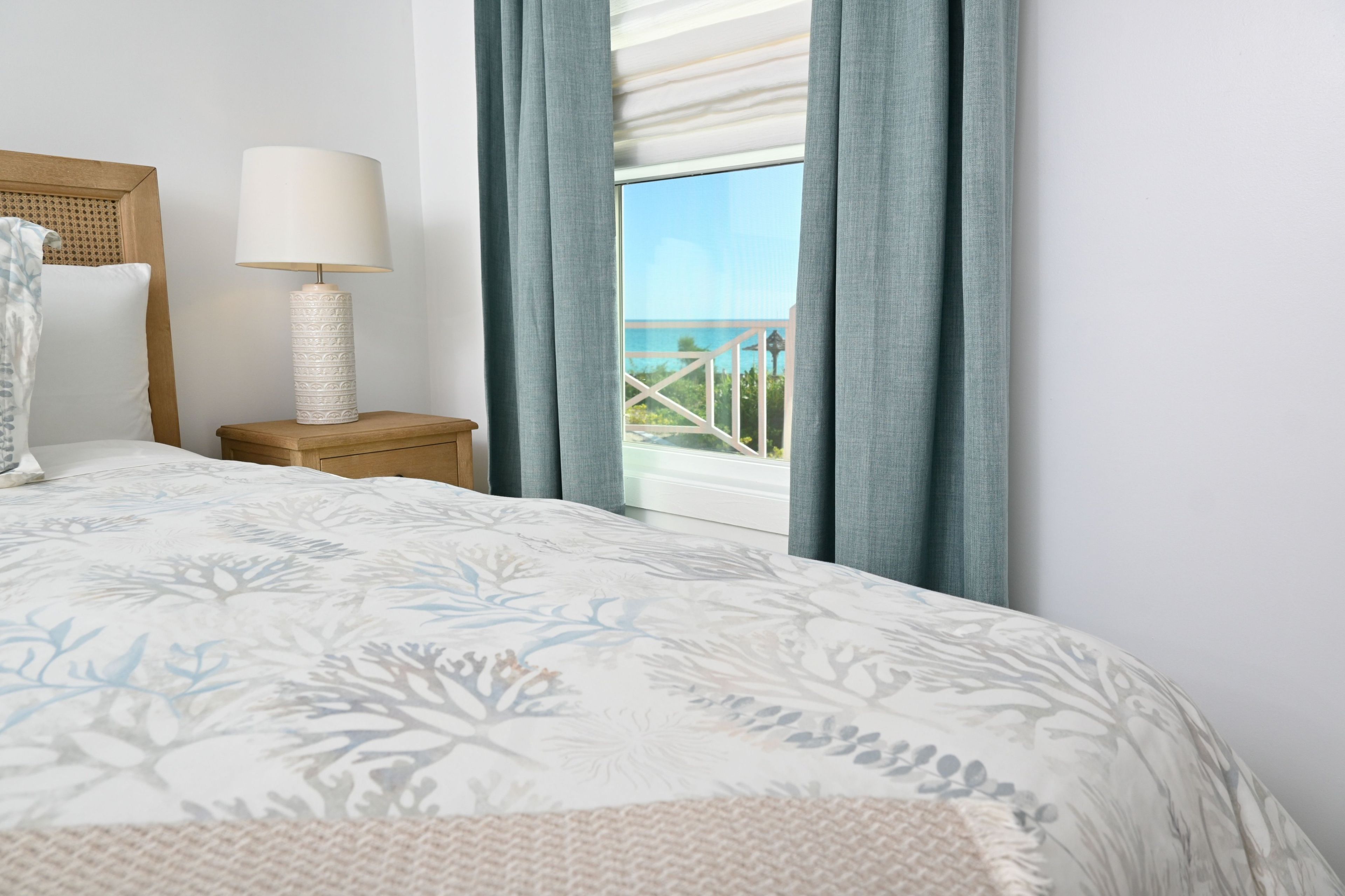 A bedroom with a bed, a bedside lamp, a nightstand, and a view of the sea.