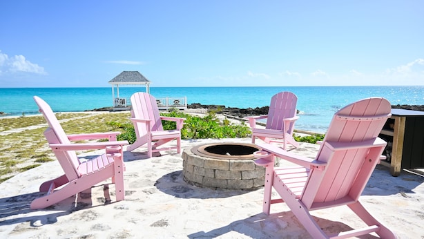 A beach scene with pink Adirondack chairs arranged around a stone fire pit, overlooking a clear blue ocean and a gazebo in the background. - Leisure Beach Ocean Retreat - Luxury Beachfront House 3 Bedroom Home (Current)