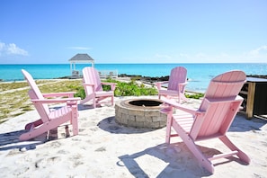 En strandscene med pink Adirondack-stole placeret omkring et ildsted i sten, med udsigt over et klart, blåt hav og en pavillon i baggrunden.