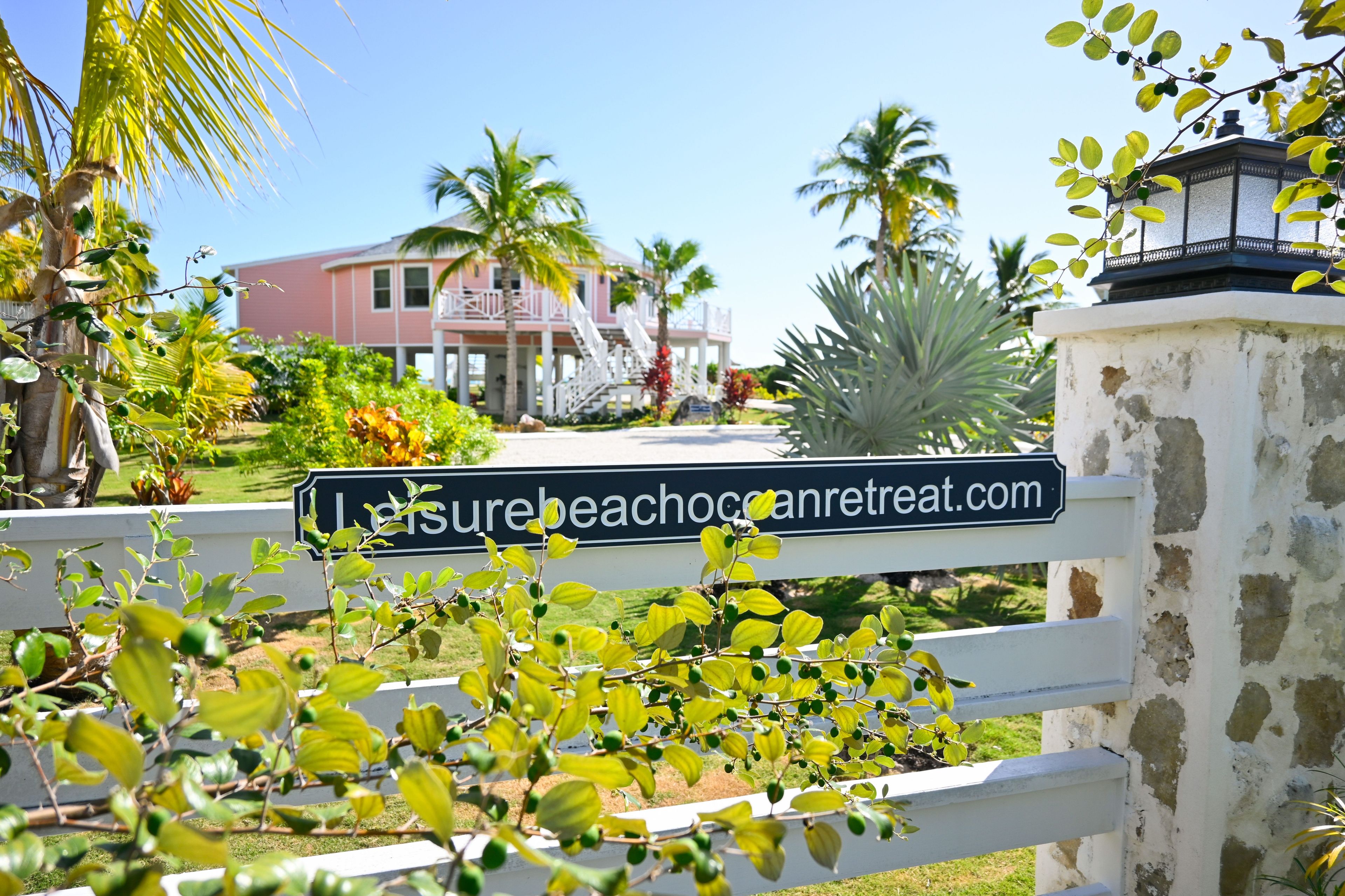 A sign with the text 'Laysurebeachcancaretreat.com' in front of a building with a white fence and greenery.