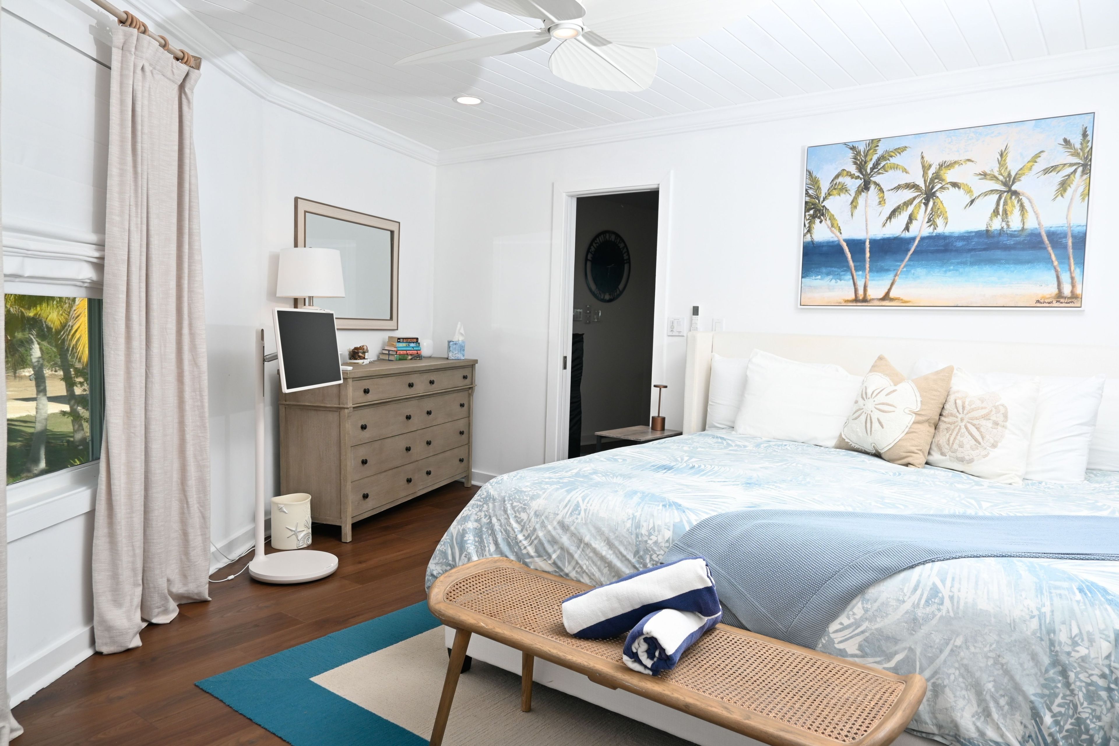 A hotel room with a bed, a desk, a chair, a window with curtains, a painting of a beach, and a ceiling fan.