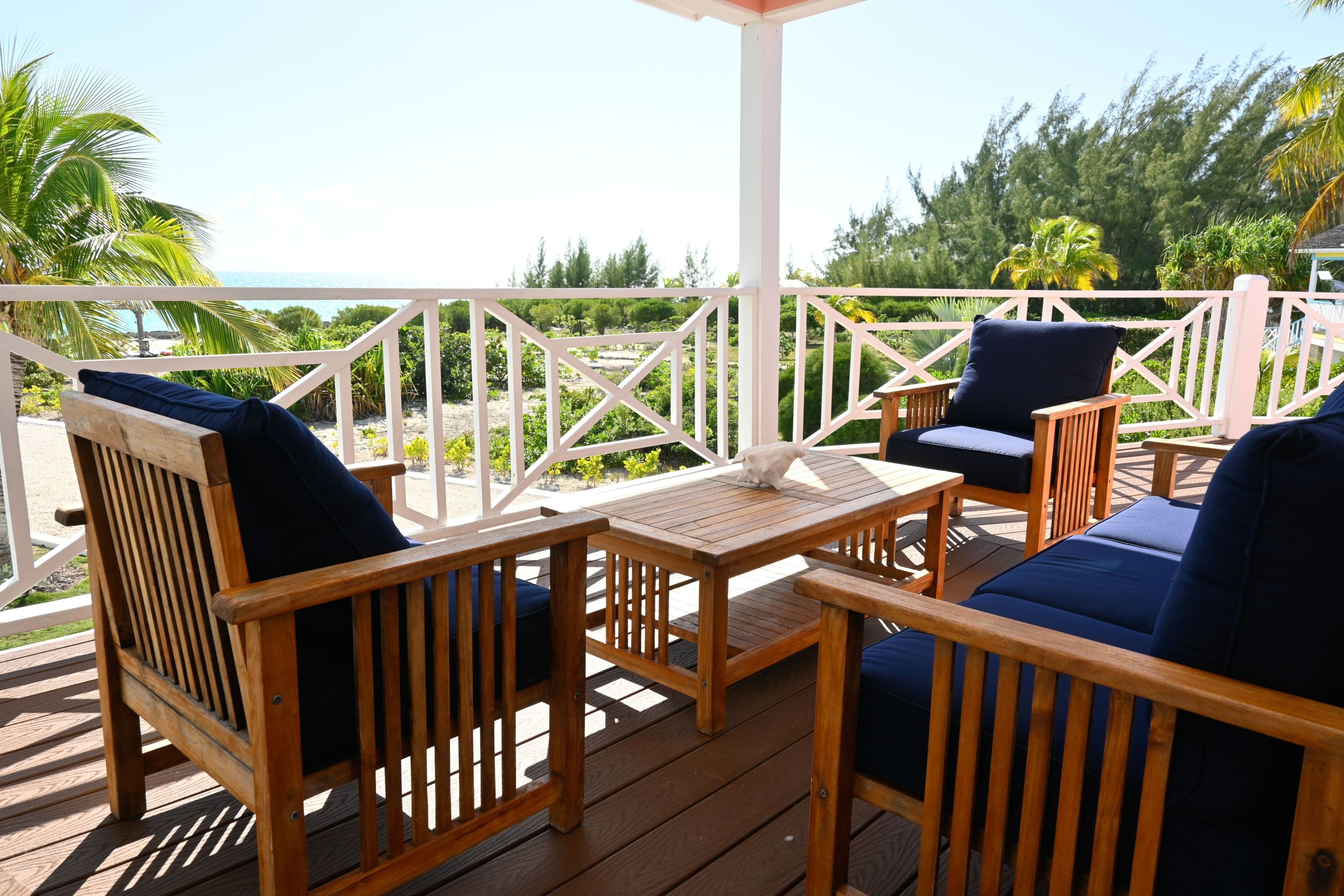A wooden deck with outdoor furniture, including chairs and a table, overlooking a beach and greenery.