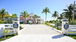 A resort entrance with a paved driveway leading to a building with a balcony, surrounded by palm trees and a clear sky. - Leisure Beach Ocean Retreat - Luxury Beachfront House 3 Bedroom Home (Current)
