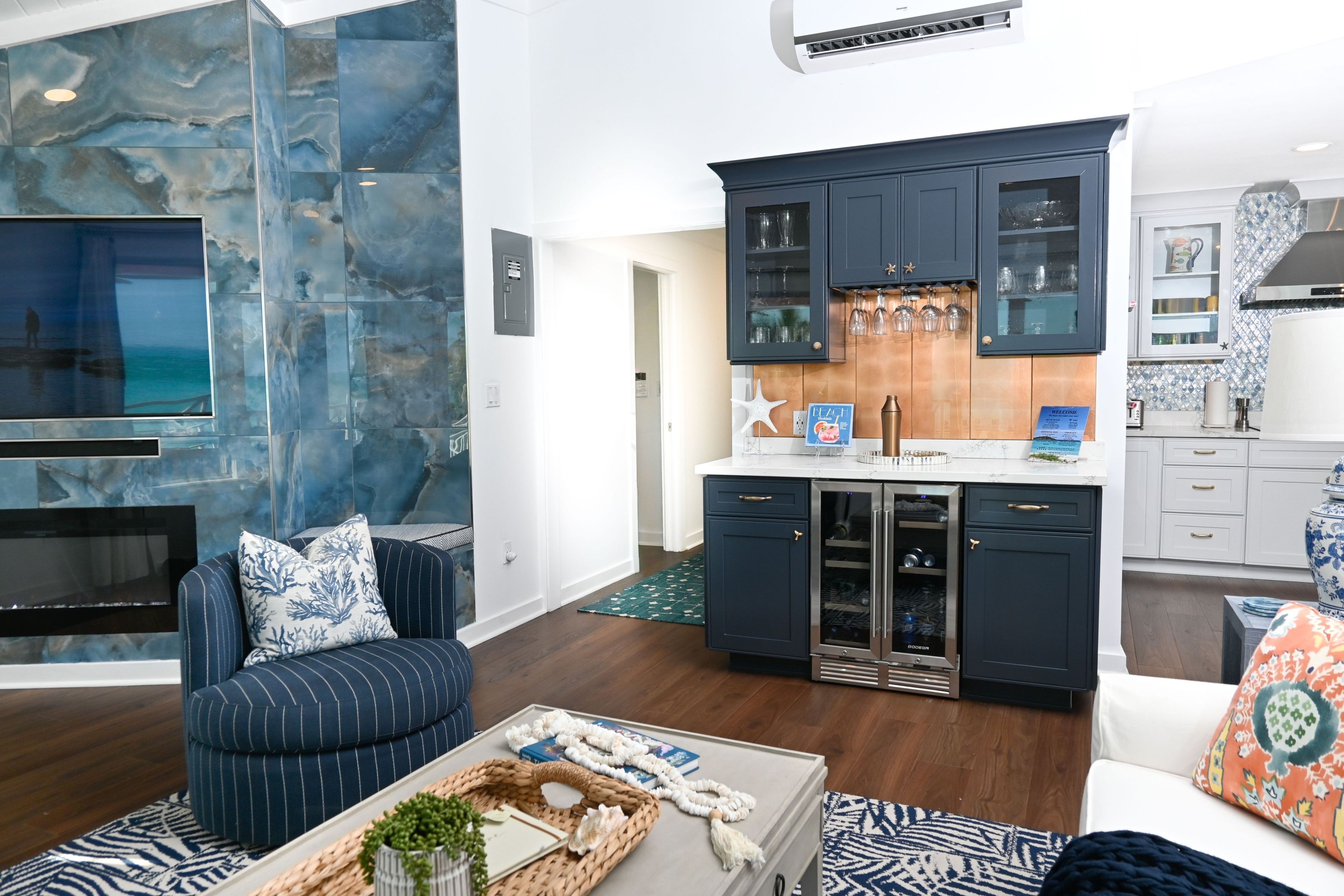 A modern living room with a large flat-screen TV, a dark blue sofa with patterned cushions, a coffee table with decorative items, and a kitchen area with dark blue cabinets and stainless steel appliances.