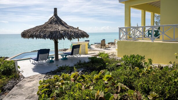Ein Strandhotel mit einem Reetdach-Sonnenschirm, Liegestühlen, einem Pool und einem gelben Gebäude mit Balkon.