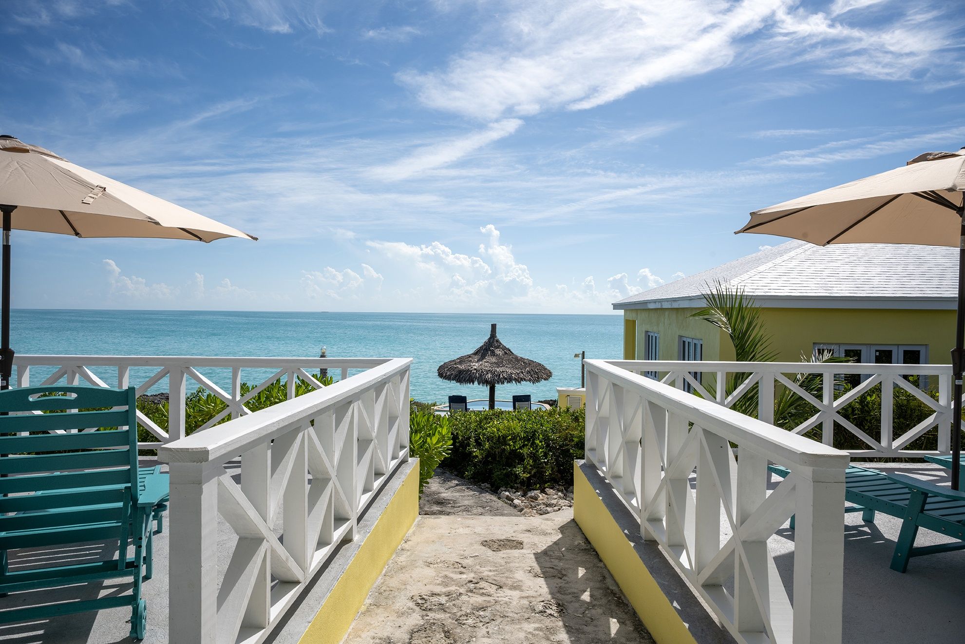 A pathway leading to a beach with umbrellas and chairs, a pool, and a view of the ocean.