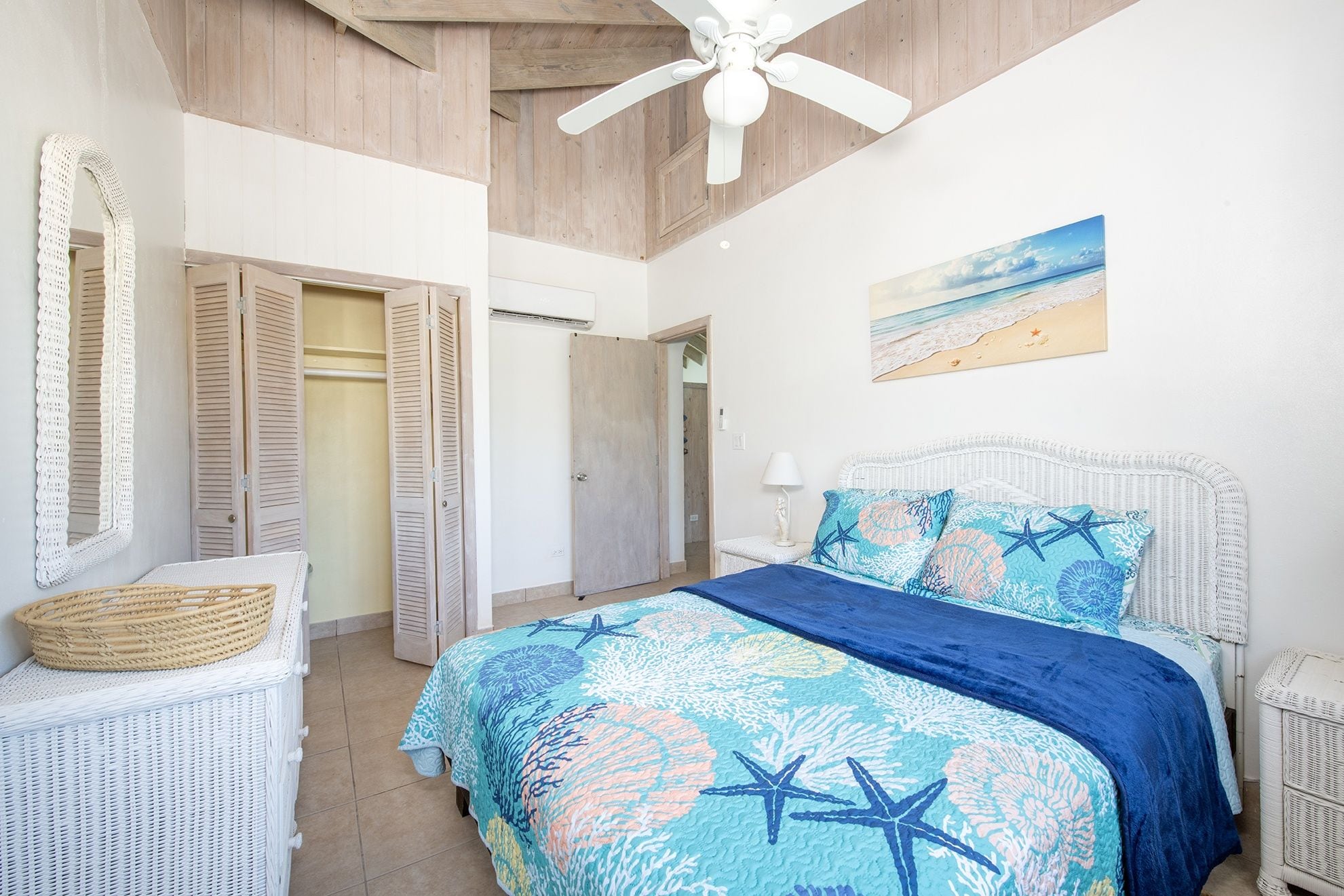 A bedroom with a bed, a ceiling fan, a beach-themed painting, and a wicker chair.