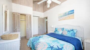 A bedroom with a bed, a ceiling fan, a beach-themed painting, and a wicker chair.