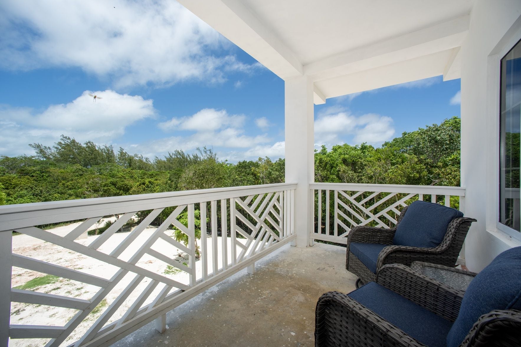 A covered outdoor area with a white railing, wicker furniture, and a view of the beach and greenery.