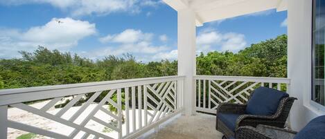 A covered outdoor area with a white railing, wicker furniture, and a view of the beach and greenery.
