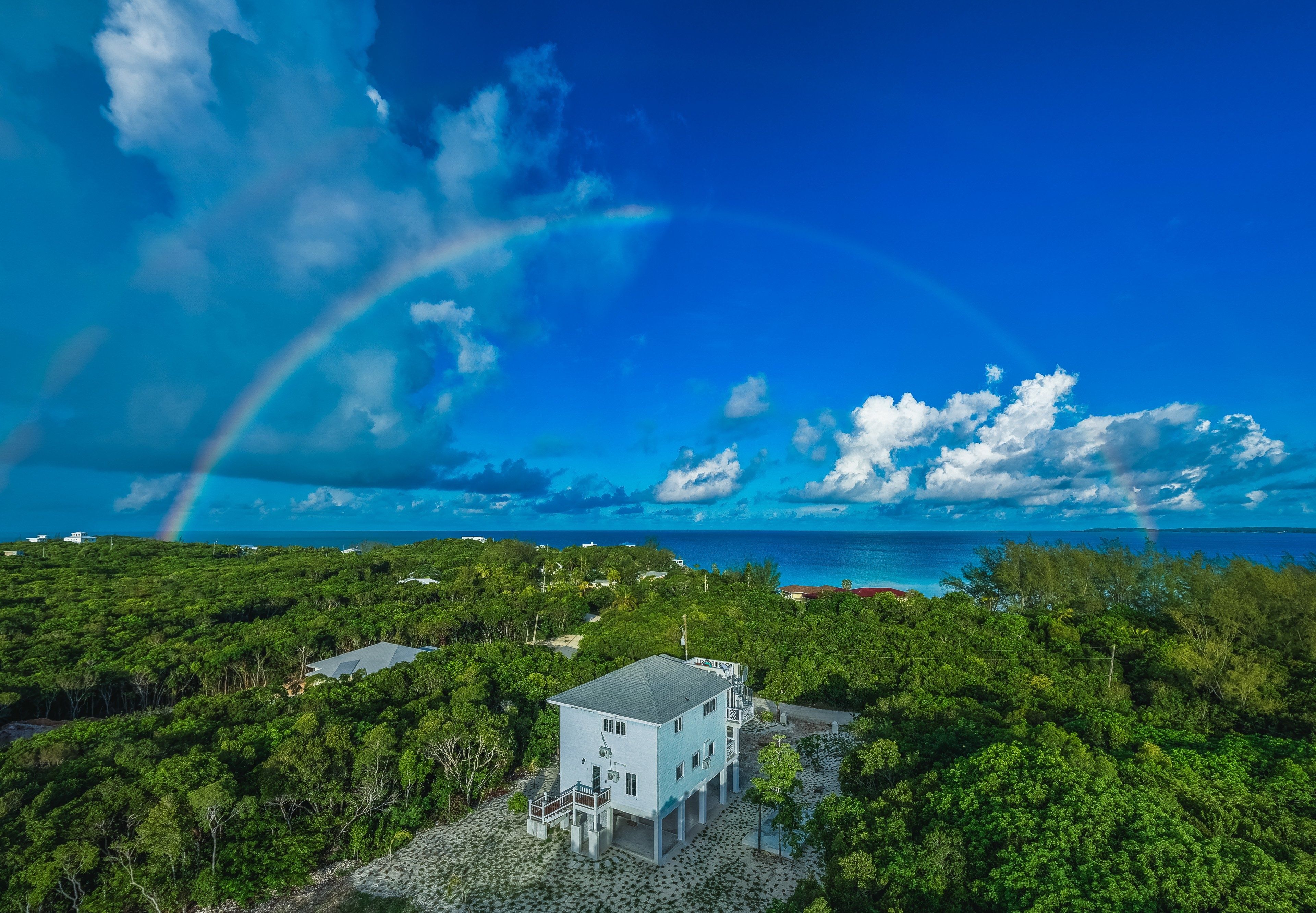 A house surrounded by lush greenery with a clear view of the ocean and a rainbow in the sky.