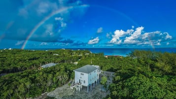 A house surrounded by lush greenery with a clear view of the ocean and a rainbow in the sky.