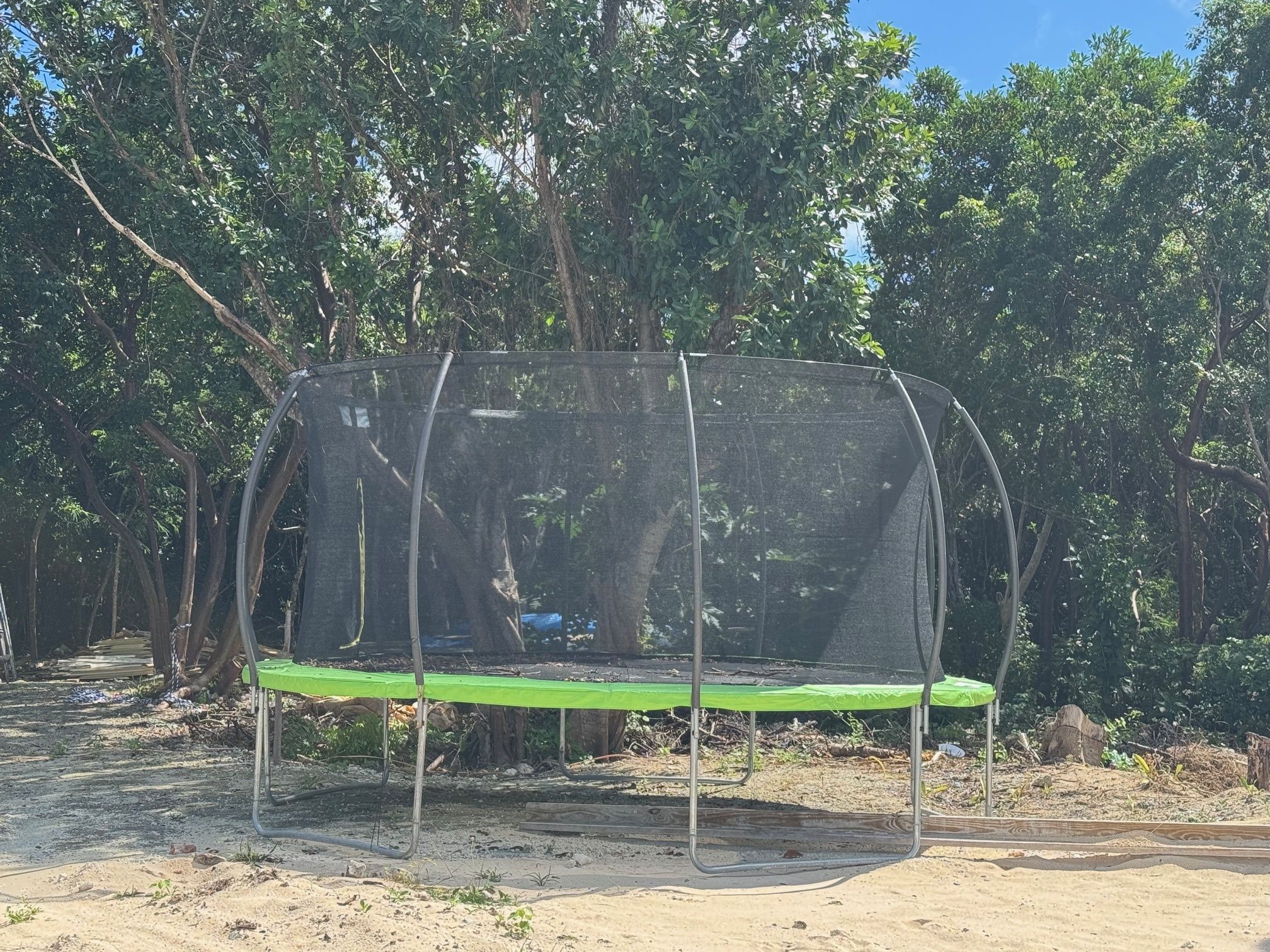A trampoline with a protective net enclosure, surrounded by trees and a dirt ground.