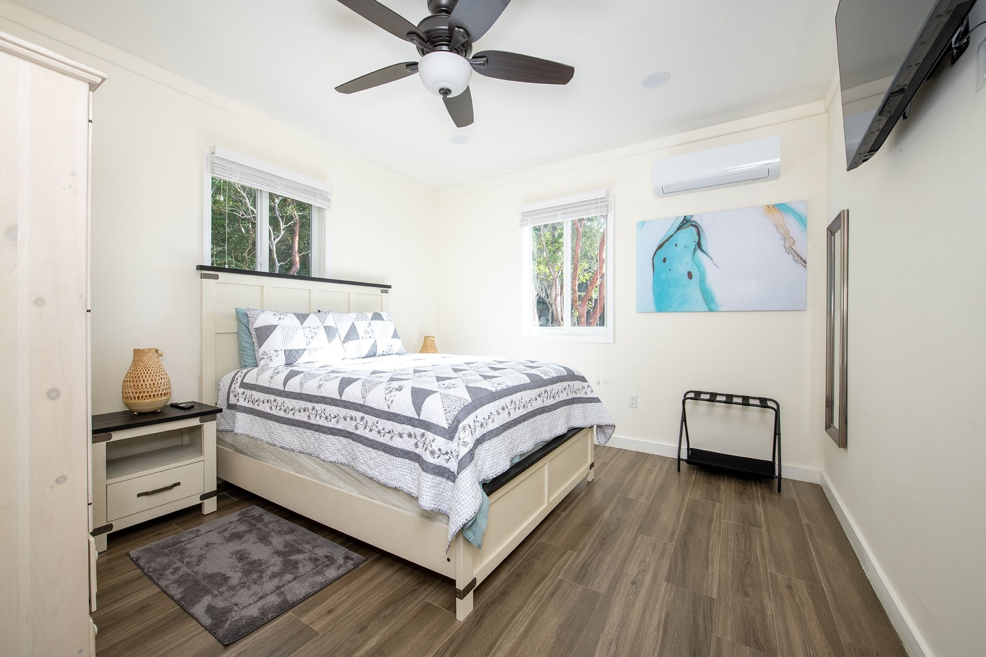 A bedroom with a bed, a ceiling fan, a window, a painting, and a television on the wall.