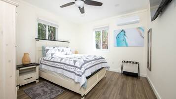 A bedroom with a bed, a ceiling fan, a window, a painting, and a television on the wall.