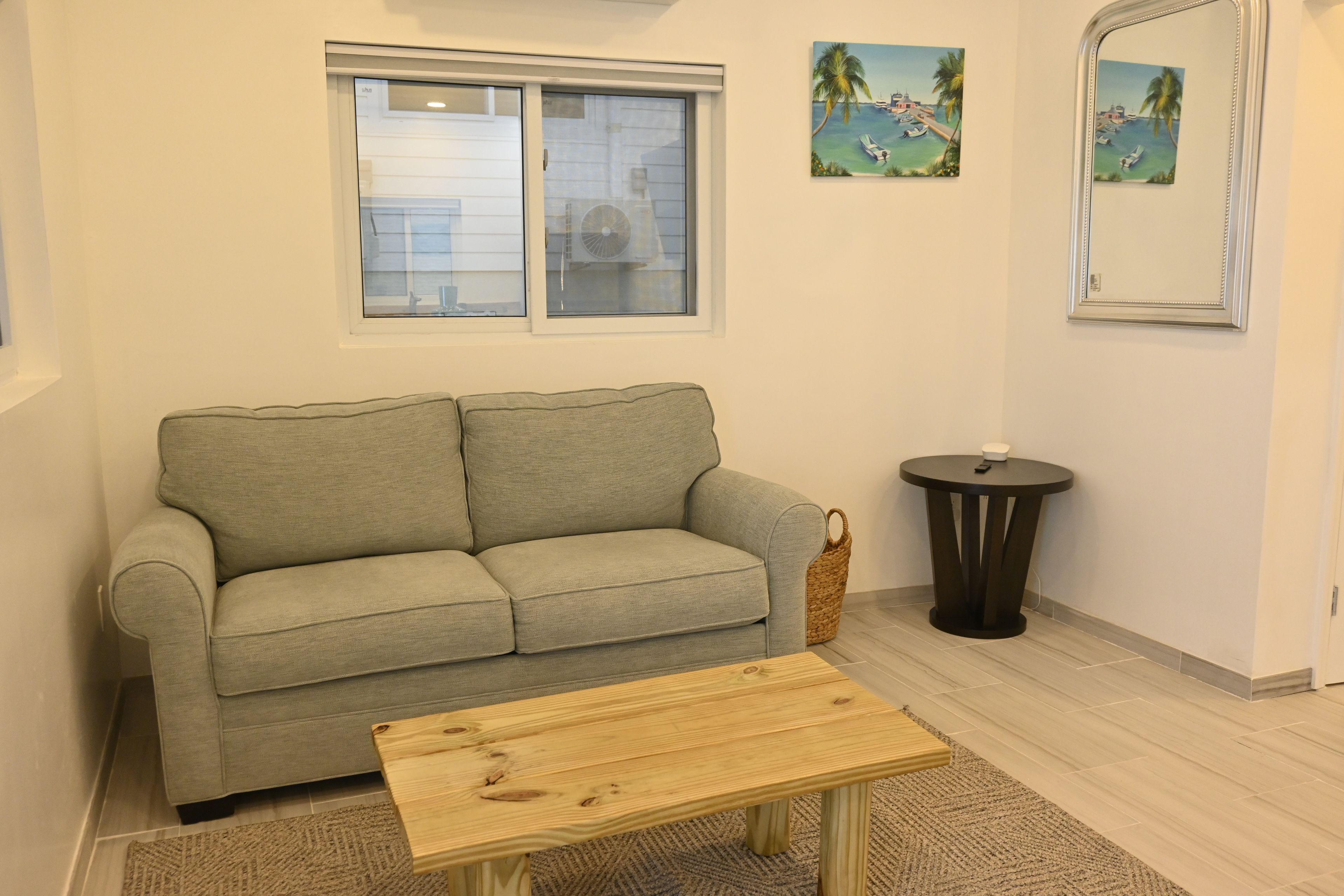 A living room with a sofa, a small wooden table, a round side table, and a window with a view of a building.
