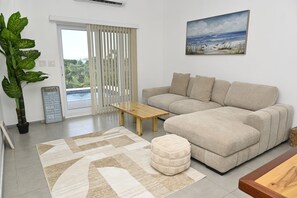 A living room with a beige sofa, a wooden coffee table, a large potted plant, a rug, and a painting of a beach scene. - Sapphire Cottage - Brand New Beach Villa With Pool 2 Bedroom Home (North Palmetto Point)