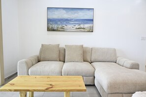 A living room with a beige sofa, a wooden coffee table, and a painting of a beach scene on the wall. - Sapphire Cottage - Brand New Beach Villa With Pool 2 Bedroom Home (North Palmetto Point)