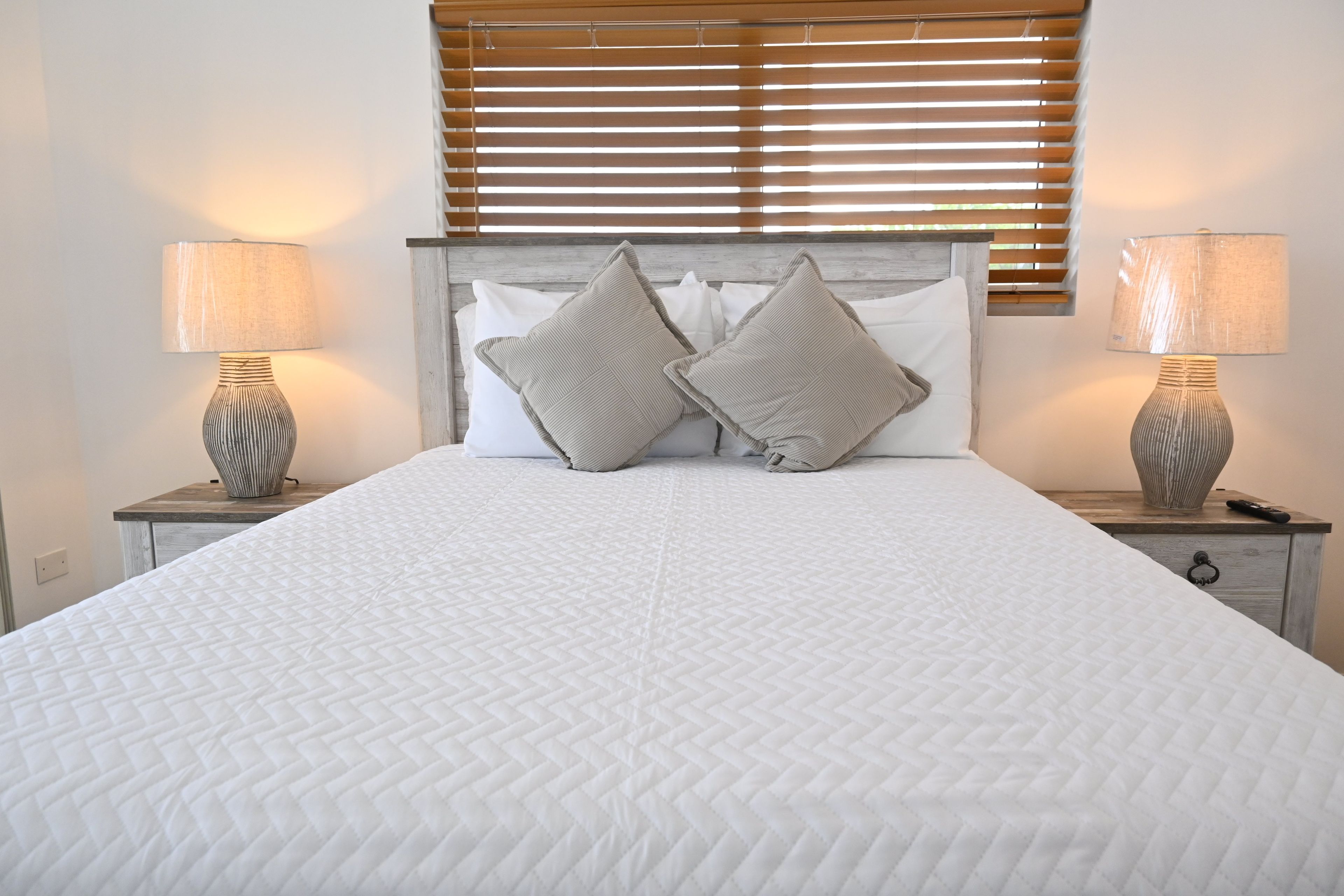 Una cama bien hecha con una colcha blanca acolchada, flanqueada por dos mesitas de noche con lámparas, y un cabecero de madera con listones horizontales.