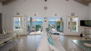 A spacious living area with a large window overlooking the sea, a sofa, a dining table, and a television.