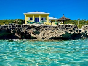 Un bâtiment jaune surmonté d’un toit de chaume, situé sur un promontoire rocheux, avec une eau turquoise et limpide en premier plan.