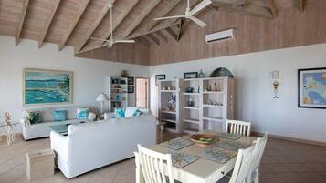 A spacious living area with a high ceiling, a dining table with chairs, a sofa with cushions, and a bookshelf.